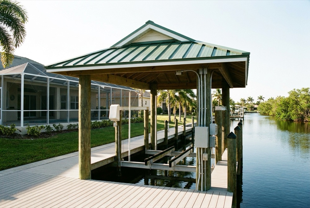 Covered boat house with a green standing-seam metal roof on timber piles, an aluminum boat lift and control boxes beneath, beside a light composite deck and quiet canal
