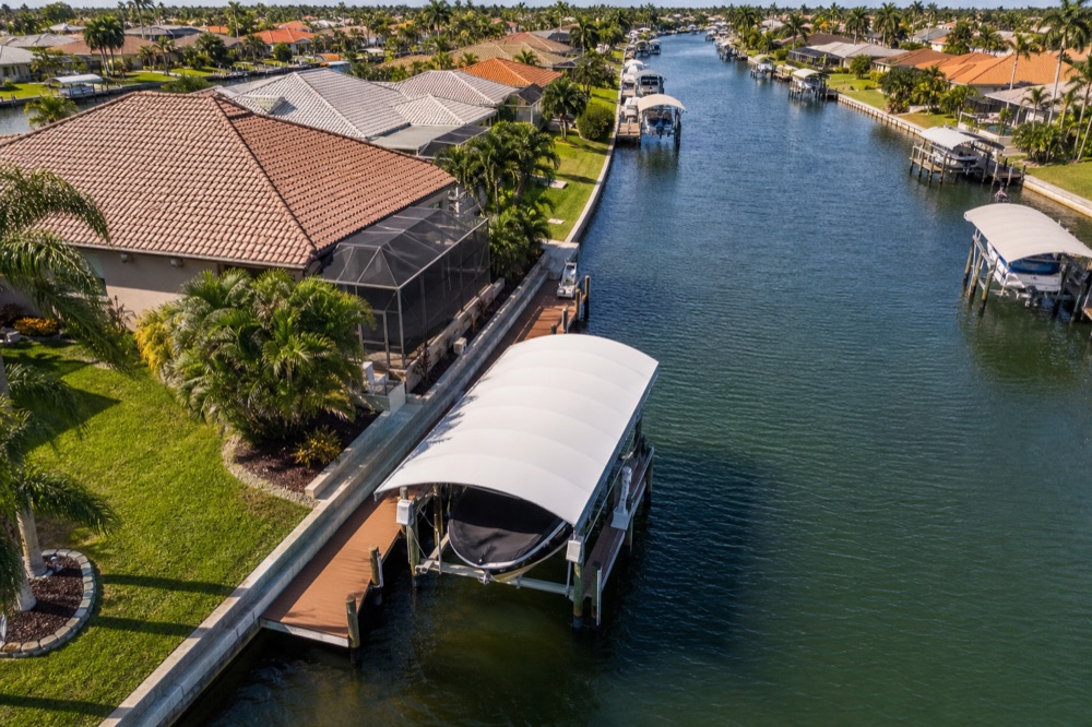 Aerial view of a canopied boat lift holding a boat alongside a composite dock, with more canopied lifts and tile-roof homes running down the canal