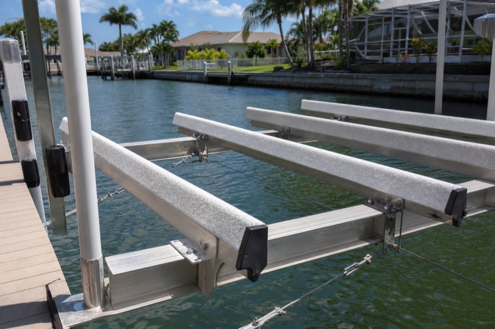 Close-up of an empty aluminum boat lift cradle showing carpeted bunks, I-beam runners, cable connections and black bumper guards over canal water