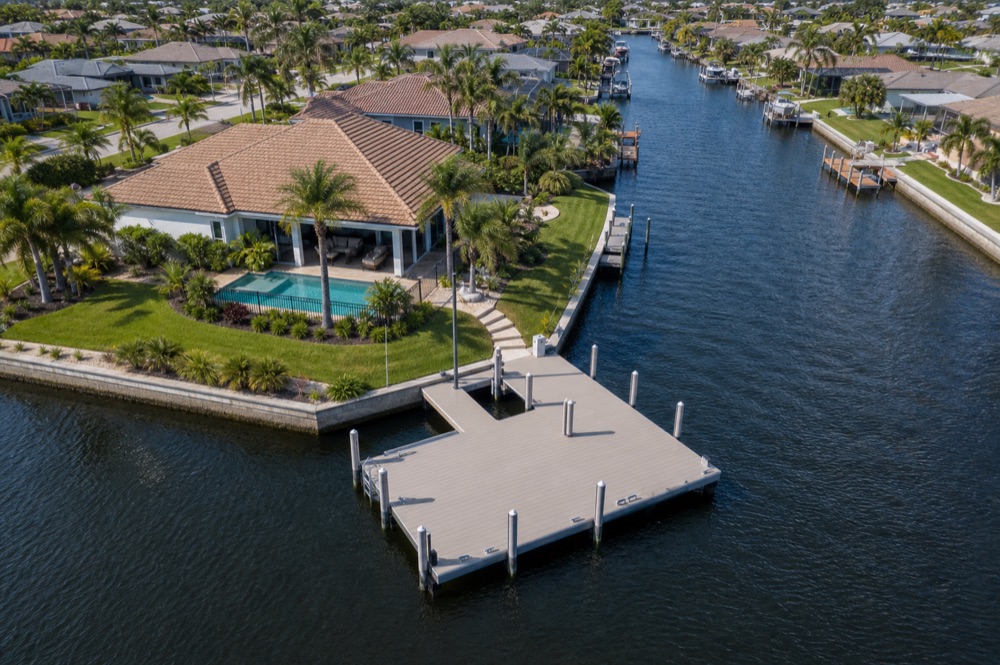 Aerial view of a large composite platform dock built off a canal point beside a tile-roof home with a pool, a concrete seawall wrapping the shoreline