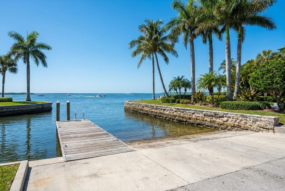 Broad concrete boat ramp sloping into shallow water beside a small timber dock and a rough limestone block seawall, with royal palms and open bay water beyond