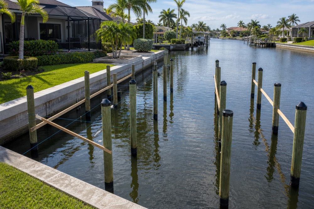 A poured concrete seawall running along a mowed lawn with newly driven pilings standing in the canal, braced with temporary wood and string lines
