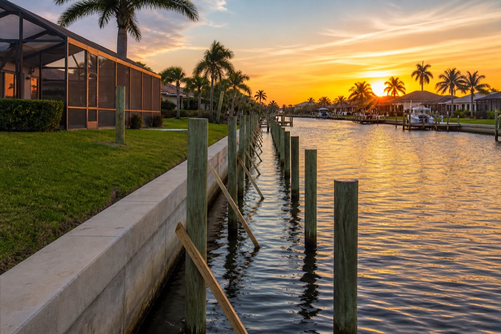 A concrete seawall and a braced row of newly driven pilings along a Southwest Florida canal at sunset, with boats and homes down the waterway