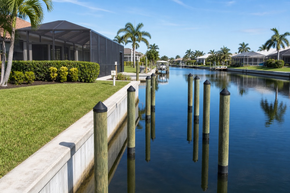 A poured concrete seawall cap running along a mowed lawn with newly driven pilings standing in the canal beyond and a screened lanai home at the left