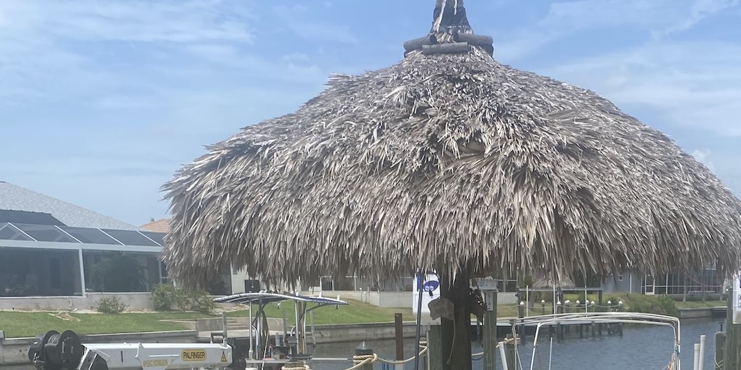 The weathered palm-thatch roof of a waterfront tiki hut, thinning and grey at the peak, with a screened lanai home across the canal behind it